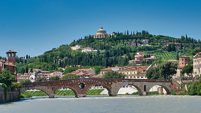 Auf einem Hügel mit Zypressen liegt die Kirche Santuario Madonna di Lourdes in Verona. Im Vordergrund alte Rundbogenbrücke über die Etsch.